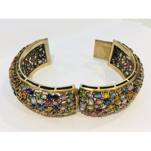 201 - A 9ct yellow gold wide hinged bangle, encrusted with a variety of multi coloured gemstones including... 