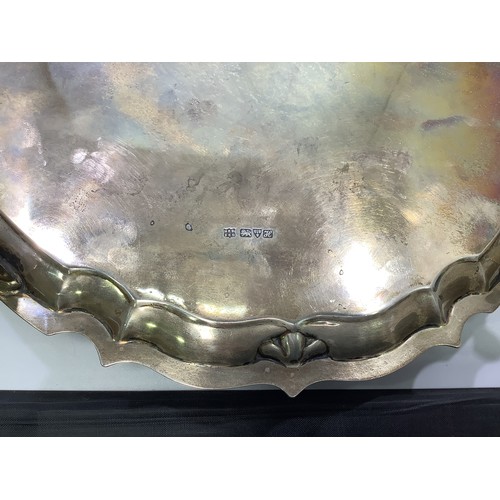 54 - A circular silver tray with shaped rim, foliate engraving and central ‘R’, raised on four pad feet, ... 