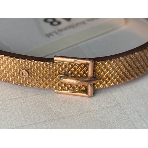 318 - A 9ct gold bangle, designed as a buckle to the top, with safety chain, weighs 10.9 grams.

The top o... 