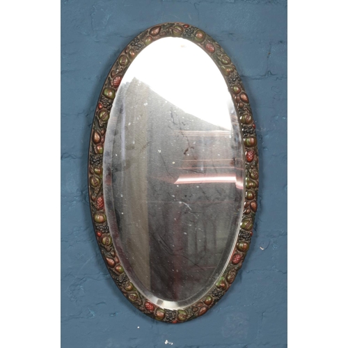 370 - A bevel edge elliptical mirror surrounded in fruit decorated frame. Approx. dimensions 67cm x 39cm.