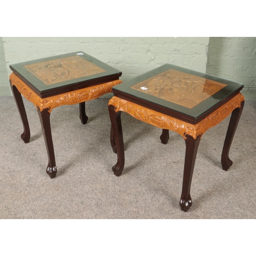 564 - A pair of Oriental hardwood occasional tables. Having deep carved decoration to tops depicting figur... 