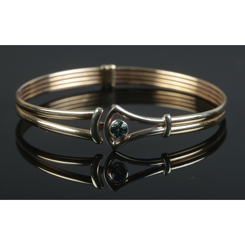 534 - A 9ct gold bangle set with a single blue topaz stone. 6.3cm at widest points. 8.64g.