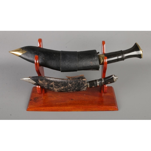 98 - Two Indian Kukri knives on display stand with larger knife featuring two small accompanying knives. ... 