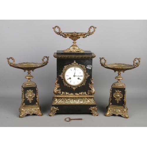 A French-style three-piece mantel clock garniture. Comprising of clock flanked by two tazza with trophy motifs, black slate base and bronze cast in a gilded Rococo style. Clock features a bayonet front bezel, white enamel dial with Roman numerals, gilt scrolling and foliate decoration and a silk suspension movement visible at the back. Clock size W25 x D13 x H45cm, tazza height 30cm.
