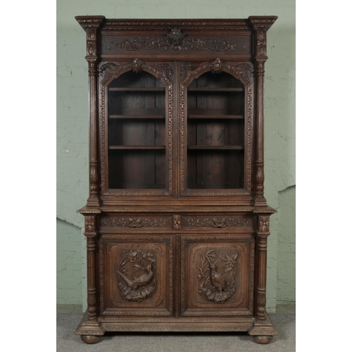 An extensively carved two section library bookcase of grand proportions. With fluted columns to either side and carved detailing throughout, a matching pair of carved pheasant panels to the lower section and original glazed panels to the top. Height 243cm, Width 150cm, Depth 60cm.