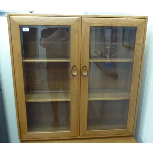 80 - An Ercol two part blonde elm cabinet, the upper part having a pair of clear bevelled glass panelled ... 