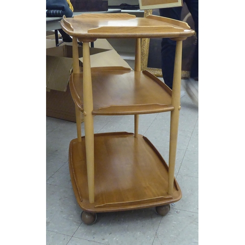 369 - An Ercol blonde beech and elm framed, three tier serving trolley, raised on turned supports and cast... 