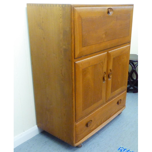 307 - A blonde Ercol elm side cabinet with a fall flap, over two cupboard doors and a long drawer, on a pl... 