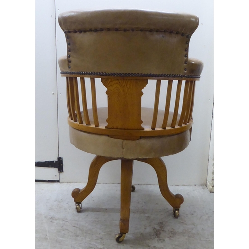 142 - An early 20thC light oak framed desk chair with a round back and square, vertical rails, upholstered... 