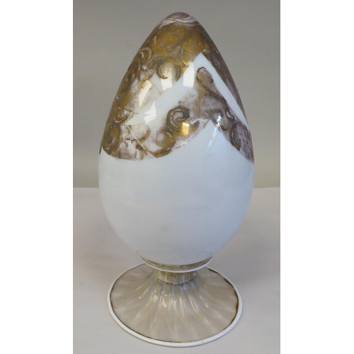 76 - An antique Murano glass two part egg ornament, overpainted and gilded with floral designs, on a milk... 