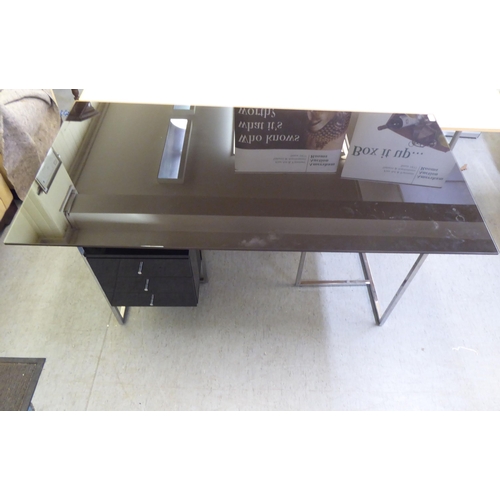 115 - A modern chrome framed, glass top desk with three drawers, raised on rectangular legs  30