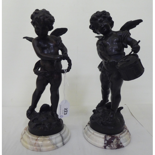 223 - A pair of bronze cherubic figures, posing with instruments, each raised on a marble plinth  bears in... 