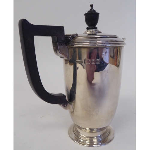 168 - A silver hot water pot of baluster form with a hinged lid, ebonised handle and knop, on a splayed fo... 