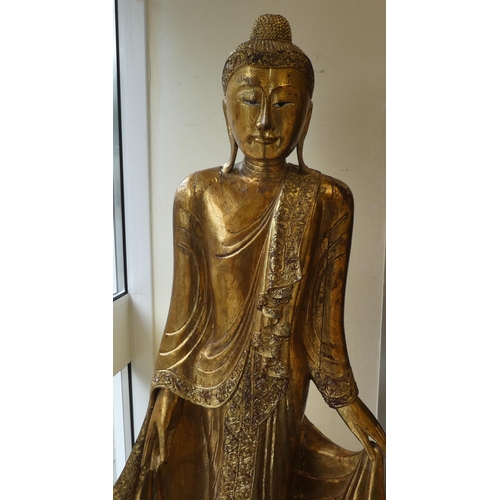 29 - A Burmese carved and gilded teak, standing temple Buddha, holding a myrobalan fruit, on a lotus flow... 