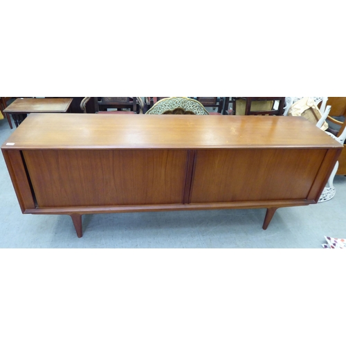 355 - A 1960/70s teak sideboard with two tambour sliding doors, enclosing a shelved interior, raised on tu... 