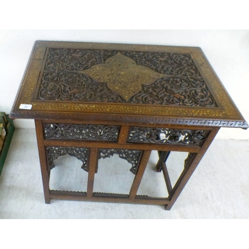 52 - An early 20thC Indian brass inlaid, carved fruitwood, folding occasional table, the drop-in top rais... 