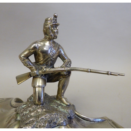 19 - An early 20thC cast white metal inkstand, surmounted by a white metal on copper military figure, kne... 