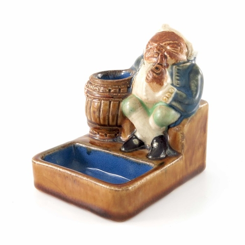 676 - Harry Simeon for Doulton Lambeth, a stoneware match striker, modelled as a seated Toby next to a bar... 