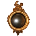 A Regency period Convex Mirror, the carved giltwood frame crested with a stylized dolphin above leaf... 