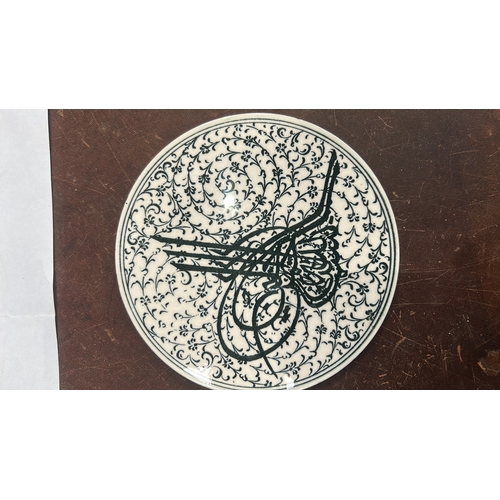 127 - Glazed ceramic round plate decorated in black with an Ottoman tughra surrounded by dense arabesque f... 