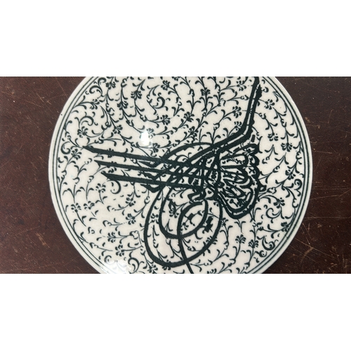 127 - Glazed ceramic round plate decorated in black with an Ottoman tughra surrounded by dense arabesque f... 
