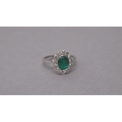 87A - An 18ct white gold ring rubover-set with an oval emerald and a halo of round brilliant cut diamonds,... 