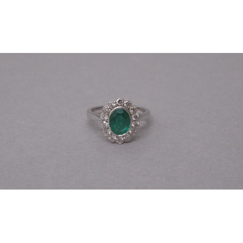 87A - An 18ct white gold ring rubover-set with an oval emerald and a halo of round brilliant cut diamonds,... 