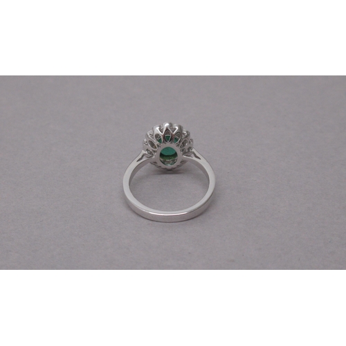 87A - An 18ct white gold ring rubover-set with an oval emerald and a halo of round brilliant cut diamonds,... 