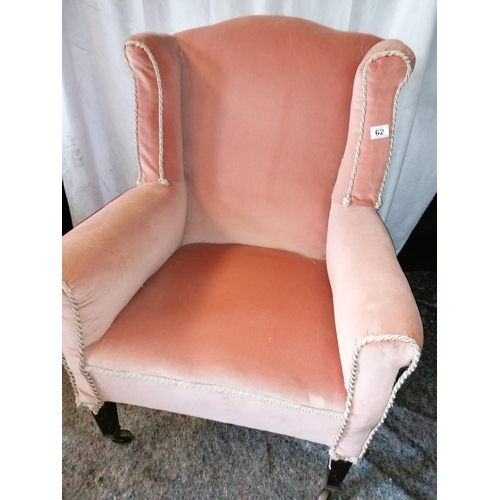 62 - An antique wing back armchair on castors, upholstered in salmon pink Draylon, cream braid piping - s... 