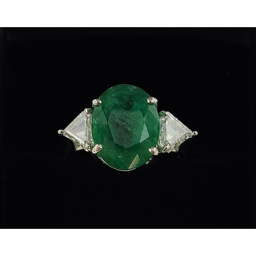 24 - AN 18CT WHITE GOLD EMERALD AND DIAMOND RING, the central oval cut emerald flanked by trilliant cut d... 