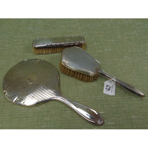 63 - Silver backed dressing table mirror, hand brush and one other brush. (3)