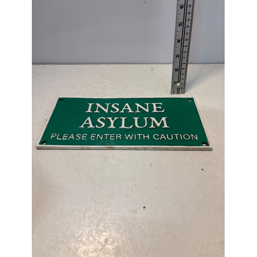 85 - Green metal sign with white text reading 'Insane Asylum: Please Enter With Caution,' featuring mount... 