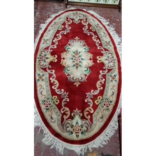 506 - Oval red floral rug with intricate beige, green, and blue designs, featuring fringed edges.