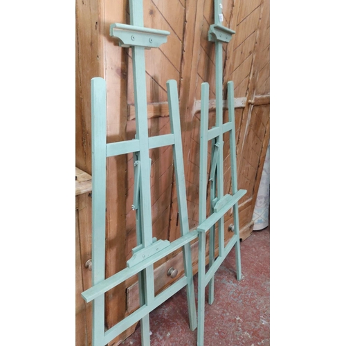 383 - Pair of adjustable wooden artist easels, painted in mint green, featuring shelf supports.