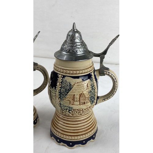 23 - Pair of ceramic steins with pewter lids, featuring a pastoral relief design. Marked 