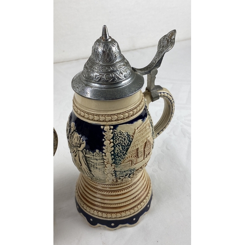 23 - Pair of ceramic steins with pewter lids, featuring a pastoral relief design. Marked 