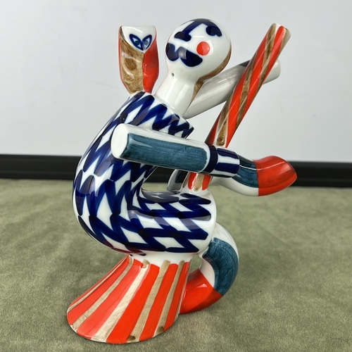 A Mid Century Sargadelos porcelain figure of a stylised witch, seated and holding a striped staff, decorated in underglaze blue with iron-red and gilt accents, raised on an integral red-striped base; factory mark to underside.