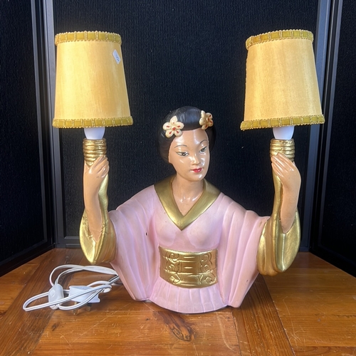 Mid-20th century figural table lamp modelled as a woman in kimono, polychrome painted and gilt, her raised arms supporting twin candle-form sockets with yellow fabric shades; wired with in-line switch.