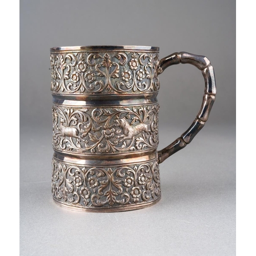 A late 19th century Kutch silver christening mug by Oomersee Mawjee / Oomersi Mawji of Bhuj, the mug of cylindrical form with three horizontal bands of repousse decorated and chased foliate scrolls, the central band further decorated with a hunting scene and shield cartouche engraved "VIVIENNE IRIS", bamboo style handle, the base stamped "OM" and "BHUJ", height 9.5cm, 6.28ozt, 195.3 grams (1)