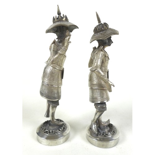 58 - A pair of Burmese silver figures, in traditional dress, holding large poles, raised on circular step... 