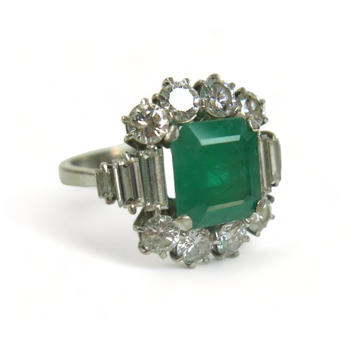 An 18ct white gold emerald and diamond ring, the central square faceted cut emerald approximately 9.2mm by 9.8mm by 5.8mm, 3.3ct, surrounded by eight brilliant cut diamonds, each 3.5mm by 2.5mm, 0.2ct, and six graduating baguette cut diamonds, London 1975, size Q, 7.8g.