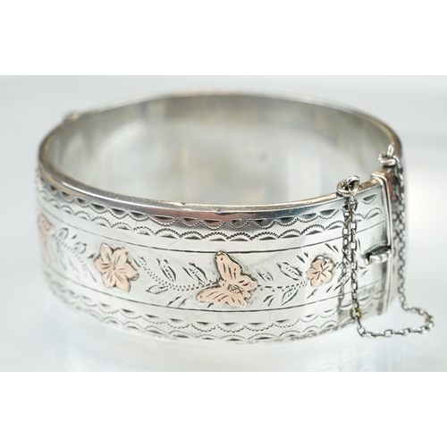 351 - Silver hinged bangle, rose gold butterfly and flower head decoration in low relief; engraved repeati... 