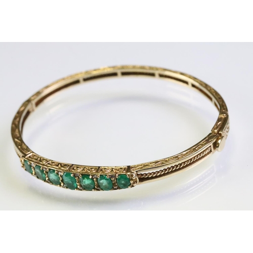 303 - Gold emerald open hinged bangle bracelet. The bracelet being set with seven oval cut emeralds with d... 