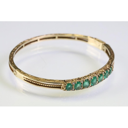 303 - Gold emerald open hinged bangle bracelet. The bracelet being set with seven oval cut emeralds with d... 