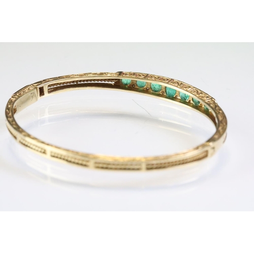 303 - Gold emerald open hinged bangle bracelet. The bracelet being set with seven oval cut emeralds with d... 