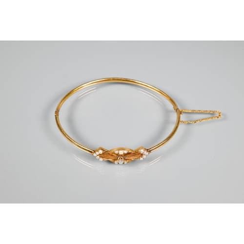 257 - A yellow gold oval half-hinged bangle of simple form with seed pearl and diamond stylised bow to cen... 