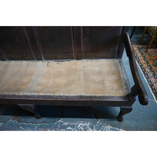 969 - An 18th century oak settle, the shaped four panel back over shaped arms and a sacking covered seat, ... 