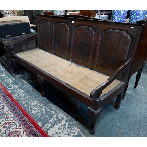 904 - An 18th century oak settle, the shaped four panel back over shaped arms and a sacking covered seat, ... 