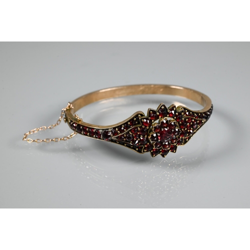 243 - A Victorian garnet-set bangle, the oval half-hinged yellow metal bangle with central flower motif, c... 