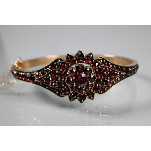 243 - A Victorian garnet-set bangle, the oval half-hinged yellow metal bangle with central flower motif, c... 